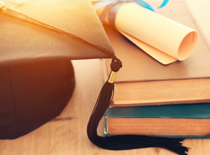 Graduation black hat over old books next to scroll diploma on wooden desk